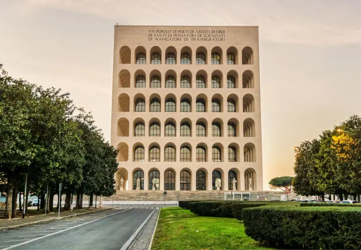 Edificio con arcate sovrapposte, simile a una stazione metro EUR Magliana, con iscrizione in latino e alberi verdi.