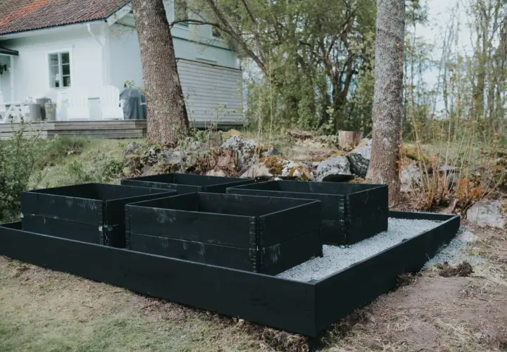 Nymålade pallkragar i svart står redo för plantering i trädgården, med ett vitt hus i bakgrunden.