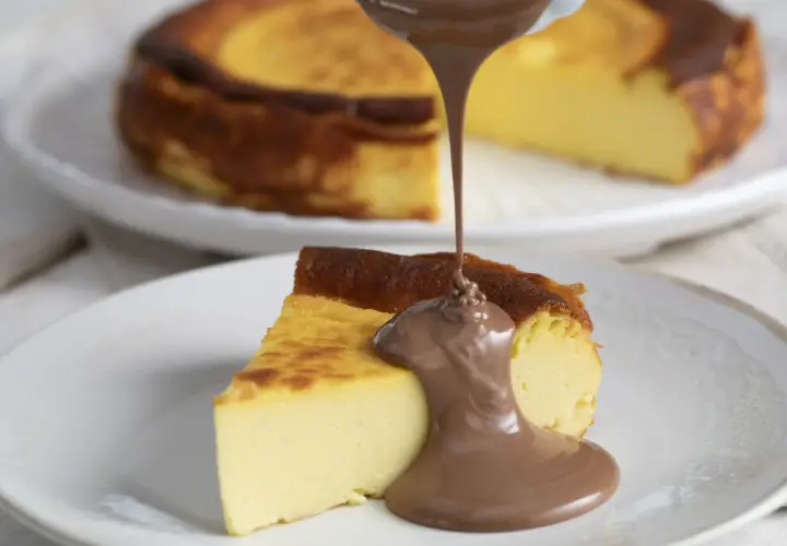 Un trozo de basque cheesecake cremoso recibe un chorro de chocolate fundido. Otro pastel entero se ve desenfocado al fondo.