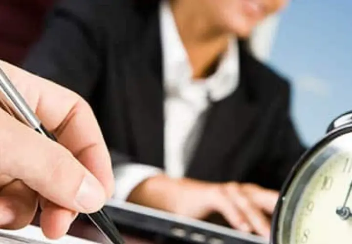 A hand holds a pen, poised to take notes, as a person in a suit works at a laptop. This scene evokes the process of creating board meeting minutes examples.