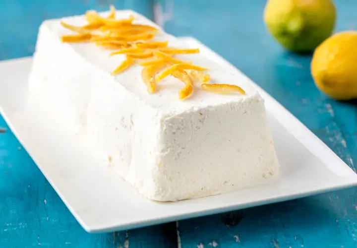 Delizioso semifreddo al limone, guarnito con scorzette candite, su un piatto bianco. Due limoni freschi sullo sfondo.
