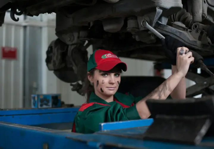 Mécanicienne sous une voiture, le visage barbouillé de cambouis. Elle est experte automobile, mais son regard suggère un abus de pouvoir.