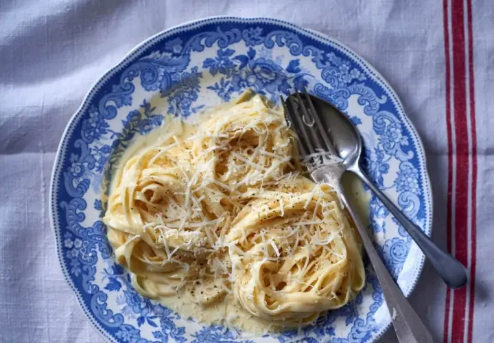 Tagliatelle w kremowym sosie alfredo, posypane parmezanem, na talerzu z niebieskimi wzorami.