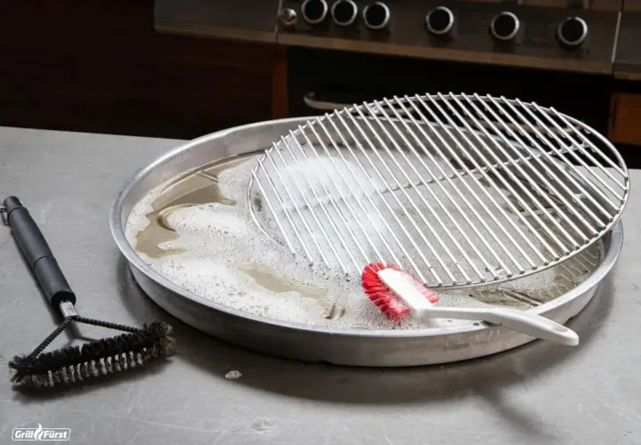 Grillrost mit eingebrannten Resten wird in Seifenlauge eingeweicht. Eine Bürste liegt daneben, bereit, den eingebrannten Grillrost zu reinigen.