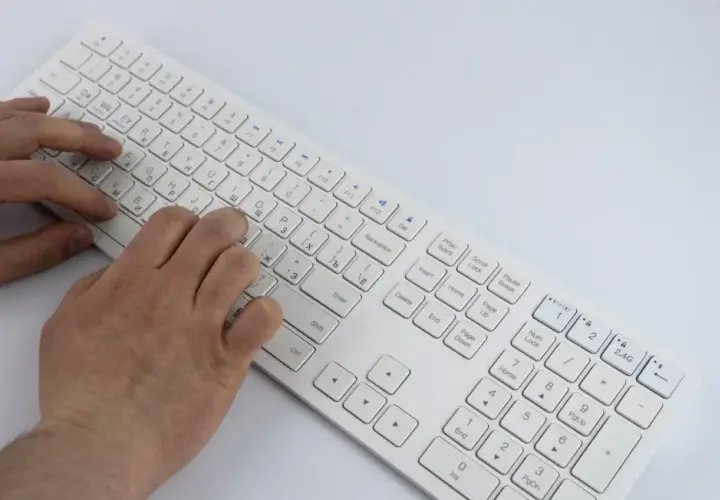 Dłonie piszą na białej klawiaturze laptopa. Widoczny jest opis klawiszy, w tym litery i symbole.