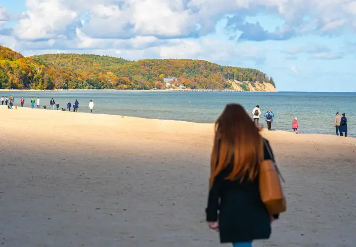 Jesień na nadmorskiej trasie spacerowej z Gdyni do Sopotu. Ludzie spacerują po piaszczystej plaży, podziwiając kolorowe drzewa na klifie.