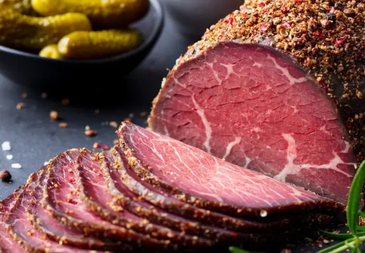 Pokrojone plastry pastrami, przyprawione pieprzem, obok ogórków konserwowych. Zastanawiasz się pastrami co to? To aromatyczne, peklowane i wędzone mięso wołowe.