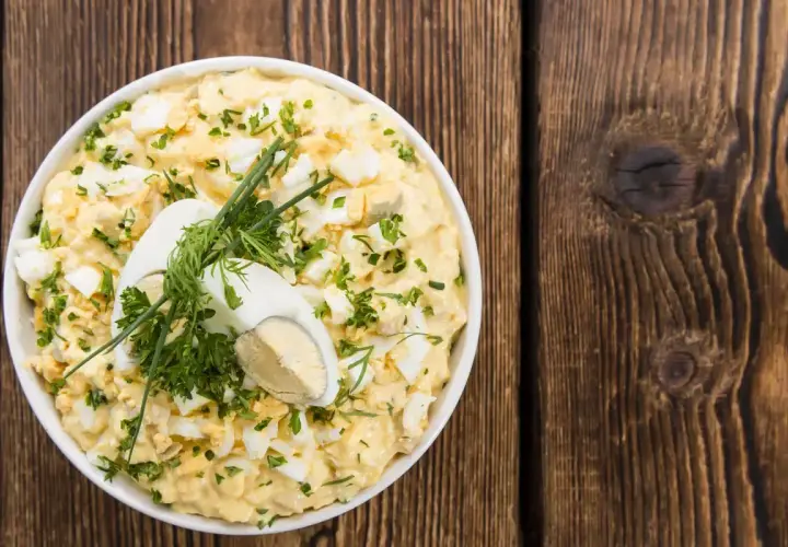 Insalata cremosa con uova di quaglia sode, erbe fresche e un uovo intero a metà come guarnizione, servita in una ciotola bianca su un tavolo di legno rustico.
