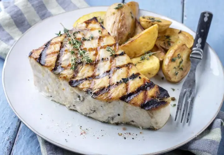 Un succulento pesce spada alla griglia, con croccanti patate arrosto e rametti di timo fresco, servito su un piatto bianco.