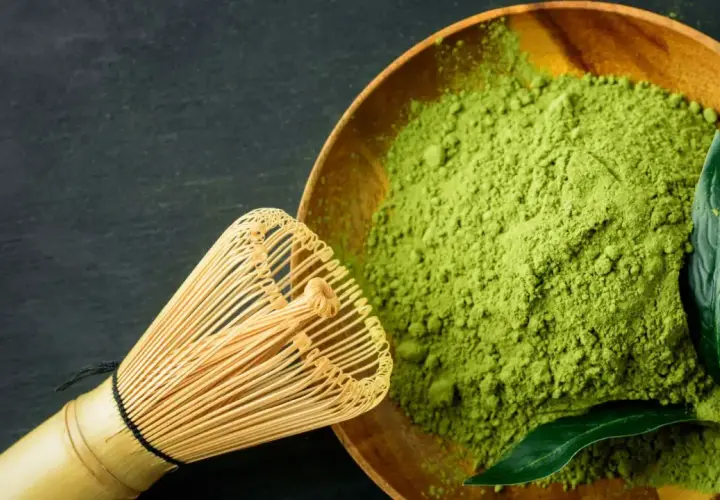 Ein hölzerner Löffel mit feinem, leuchtend grünem Matcha-Pulver und ein Bambusbesen daneben. Der Geschmack ist intensiv, leicht bitter.