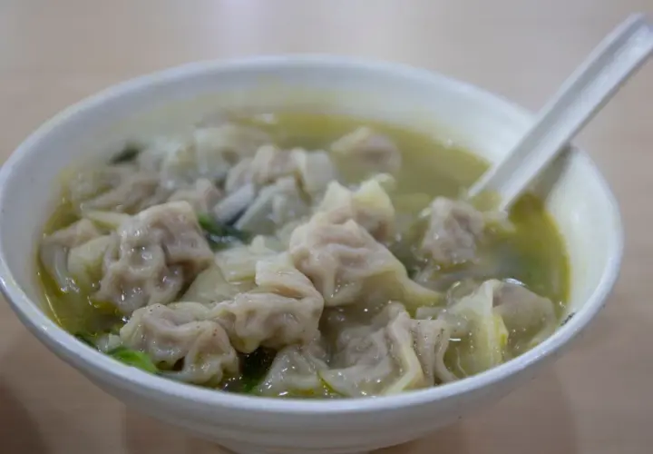 A miseczka pełna aromatycznej zupy wonton, z widocznymi pierożkami i zielonymi warzywami. Łyżka zanurzona w bulionie.