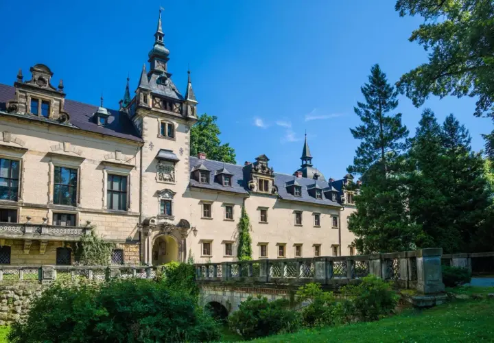Elegancki hotel zamek z wieżą i mostkiem, otoczony zielenią, pod błękitnym niebem.
