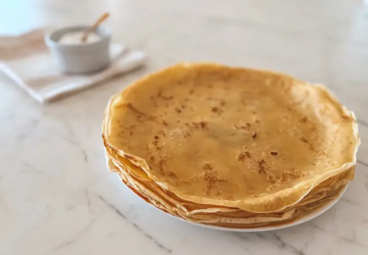 Pile de crêpes dorées, parfaites pour une recette crêpe moelleuse. Un petit bol de sucre est à côté.
