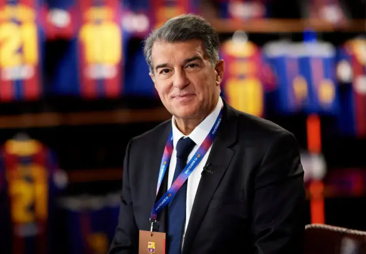 Prezes FC Barcelona, Joan Laporta, w garniturze, z identyfikatorem klubu na szyi, na tle klubowych koszulek.