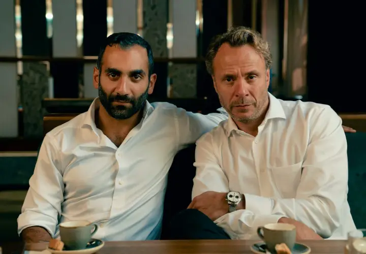Två män i vita skjortor sitter vid ett bord med kaffe. En arm vilar på den andres axel. Scen från hundarna serie.