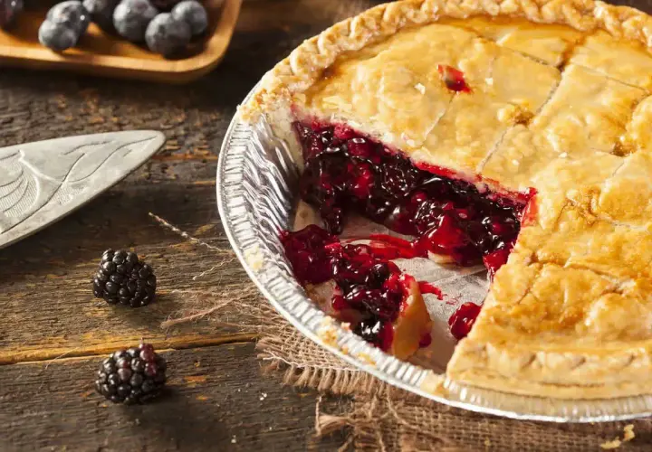 Una fetta di torta di more appena sfornata, con il ripieno succoso che cola. Due more fresche adagiate sul tavolo di legno.