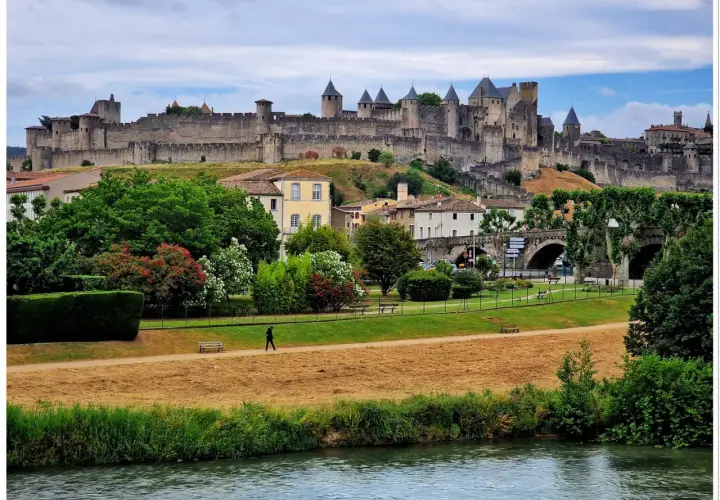 Majestatyczna twierdza Carcassonne góruje nad miastem. W dole zielony park, rzeka i samotny spacerowicz.