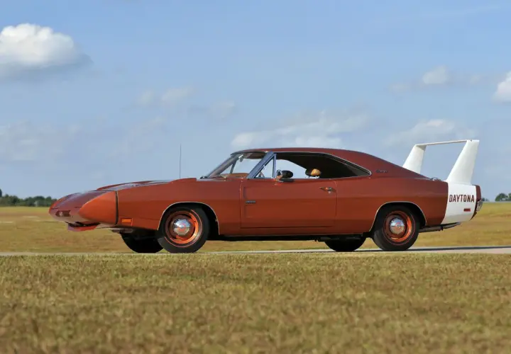 Pomarańczowy Dodge Daytona z charakterystycznym spojlerem i białym pasem z napisem "DAYTONA" na błotniku, zaparkowany na tle nieba z chmurami.