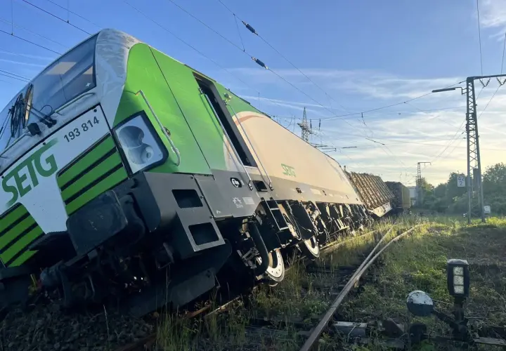 Zielony pociąg towarowy wykoleił się, przechylając się na bok na torach. W tle błękitne niebo i linie energetyczne.