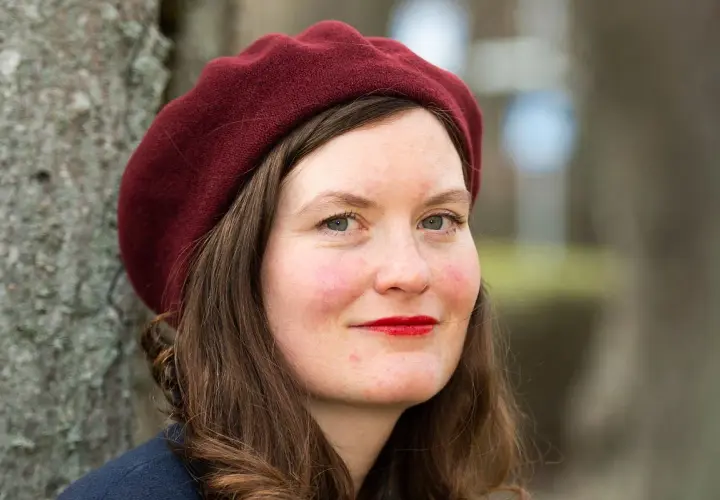 Alva Dahl, med en vinröd basker och röda läppar, ler mot kameran. Hon står utomhus vid ett träd.