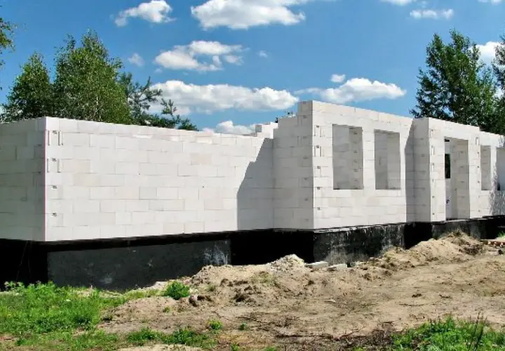 Nowe, białe ściany domu z bloczków betonowych, z otworami na okna i drzwi, stoją na czarnym fundamencie.