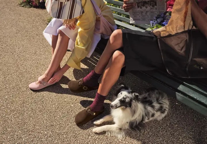 Dwie kobiety siedzą na ławce, prezentując modne buty na lato: różowe baleriny i brązowe klapki. Obok leży pies.