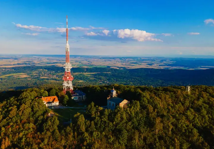 Wieża telekomunikacyjna na górze Ślęża góruje nad lasami i polami. Widać też budynki i kościółek.