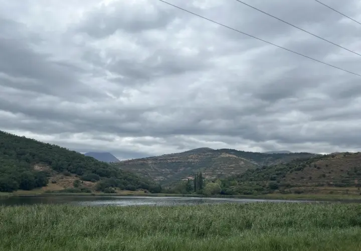 Paisaje sereno con un lago rodeado de colinas verdes y secas, bajo un cielo nublado. Las montañas a lo lejos parecen la geganta adormida.