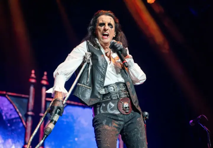 Alice Cooper na scenie z mikrofonem i kulami, w charakterystycznym makijażu i stroju.