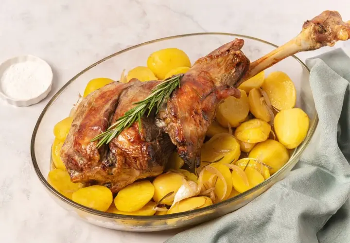 Pierna de cordero al horno dorada, servida con patatas asadas, romero y ajo en una fuente de vidrio.