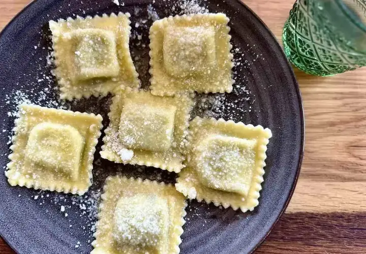 Pięć kwadratowych ravioli z ricottą, posypanych parmezanem, na ciemnym talerzu. Obok zielony kieliszek.