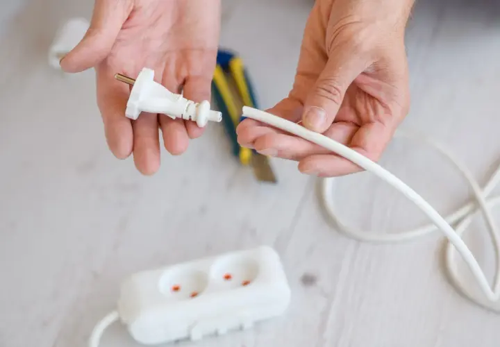 Dłonie przygotowują kabel do podłączenia do wtyczki z uziemieniem. W tle widać listwę zasilającą.