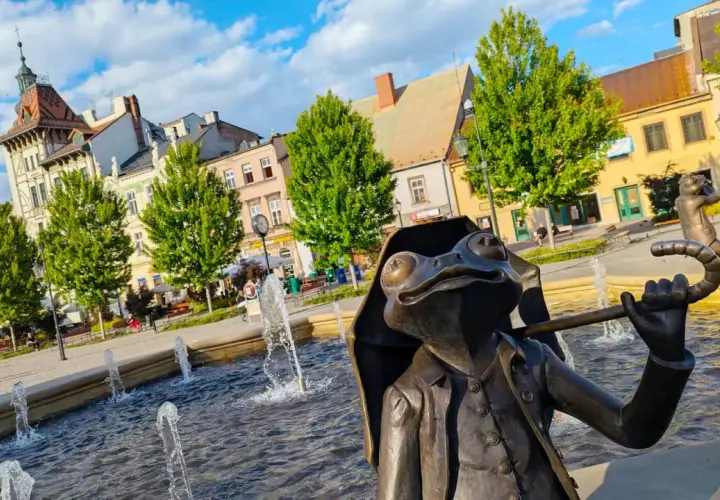 Bielsko-Biała atrakcje dla dzieci: żaba z parasolką i fontanny na rynku. W tle kolorowe kamienice i zielone drzewa.