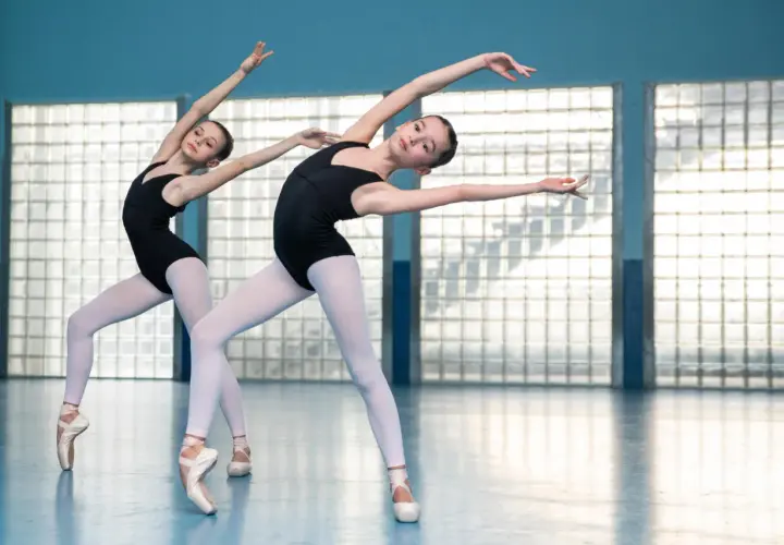 Dwie młode tancerki baletowe w czarnych trykotach i białych rajstopach, stojące na czubkach palców, wykonują elegancką choreografię.