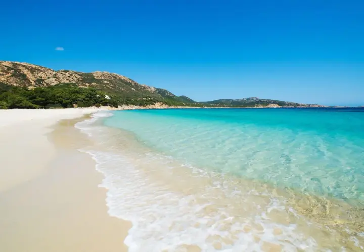 Plage de sable blanc bordée par une eau turquoise cristalline, avec des collines verdoyantes en arrière-plan. Un paysage idyllique d'**Italie bord de mer**.