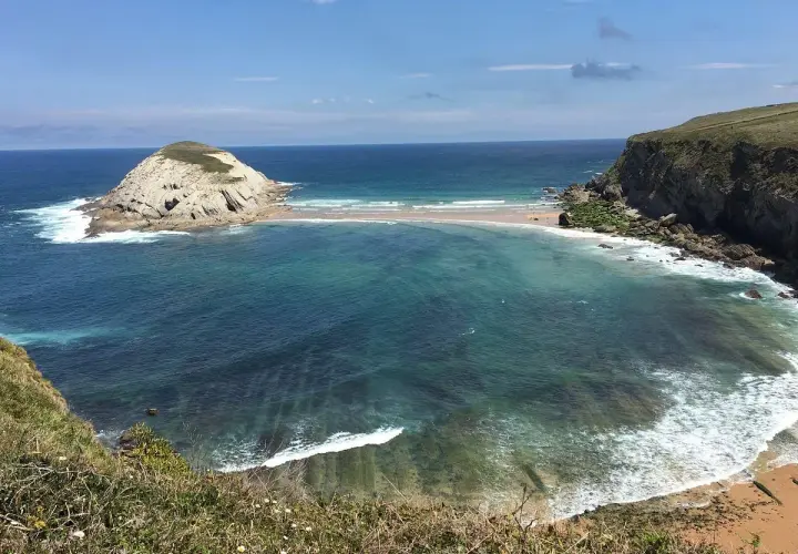 Vista de la Virgen del Mar en Santander, con una isla rocosa y aguas turquesas.