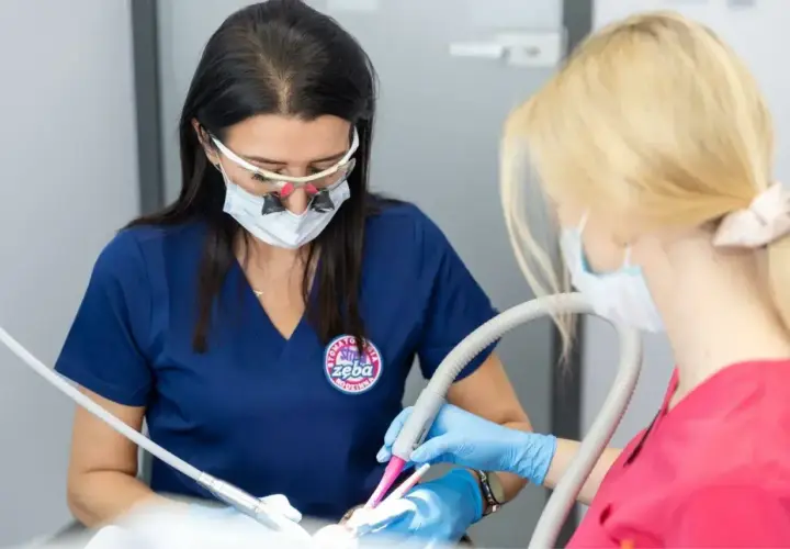 Dwie dentystki w maseczkach i okularach pracują przy pacjencie. Jedna używa wiertła, druga odsysacza. To profesjonalna artykaina.