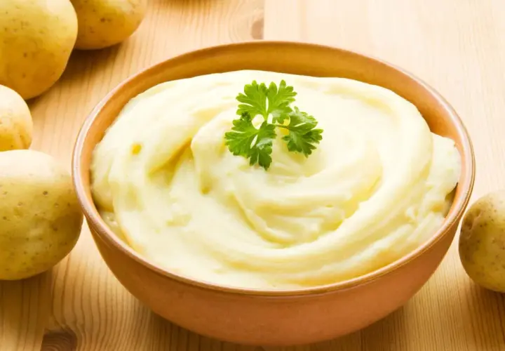 Un tazón de cremoso puré de patatas, decorado con perejil, junto a patatas crudas. ¡Así es como hacer puré de patatas perfecto!