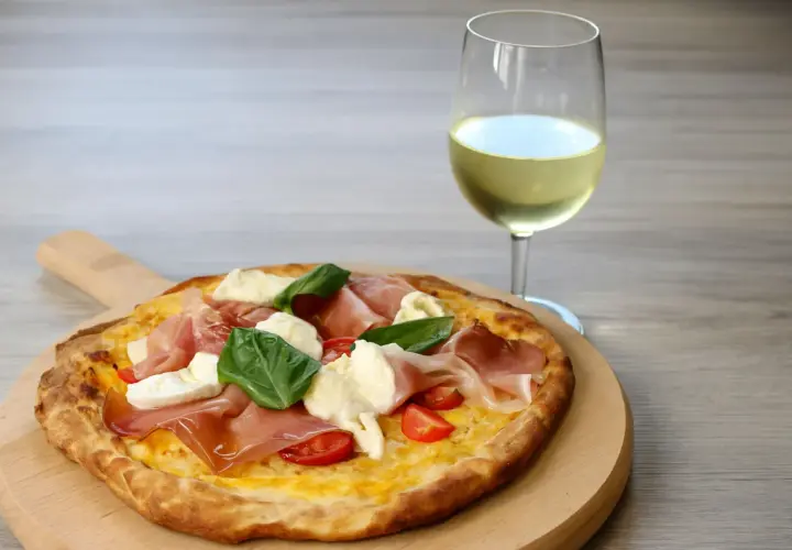 Pizza con prosciutto, mozzarella e basilico su tagliere di legno, accanto a un bicchiere di vino bianco. Un perfetto abbinamento vino pizza.