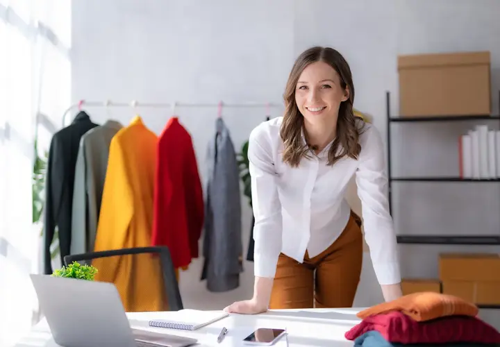 Une chef de produit textile souriante, entourée de vêtements colorés sur un portant, travaille dans son bureau lumineux.