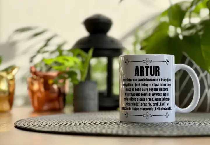 Biały kubek z napisem "ARTUR" i tekstem o znaczeniu imienia, na tle roślin.