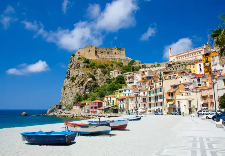 Vue d'une ville côtière italienne avec un château sur une falaise, des maisons colorées et des bateaux sur une plage.
