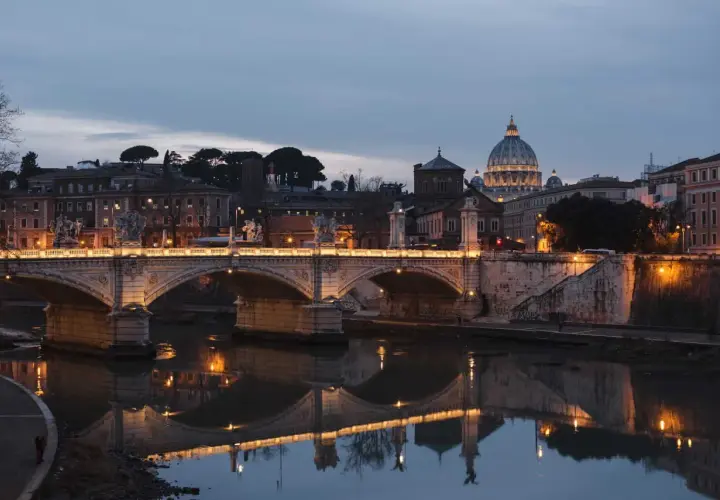 Oświetlony most Ponte Sant'Angelo nad Tybrem, z kopułą Bazyliki św. Piotra w tle. Rzeka w Rzymie odbija światła miasta.