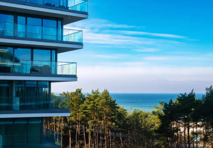 Nowoczesny budynek z balkonami i widokiem na morze. W oddali las i błękitne niebo. Idealne miejsce na wypoczynek w Wave Międzyzdroje Resort&SPA.