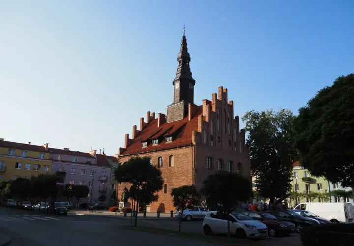 Ratusz w mieście nad jeziorem Skiertąg, z charakterystyczną wieżą zegarową, otoczony kolorowymi kamienicami i zielenią.