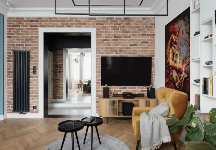 Nowoczesny salon z ceglaną ścianą, na której wisi telewizor. Oświetlenie ściany z kamienia podkreśla jej fakturę. Obok żółty fotel i obraz z gitarzystą.