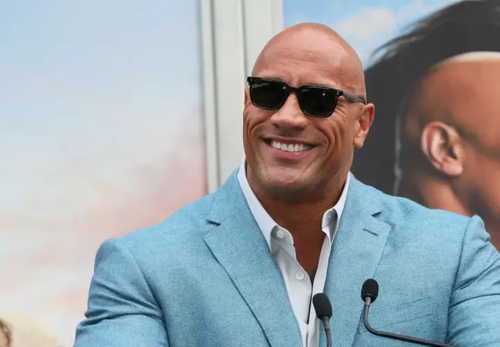 Dwayne "The Rock" Johnson, być może najbogatszy aktor na świecie, w błękitnej marynarce i okularach przeciwsłonecznych, uśmiecha się do kamery.