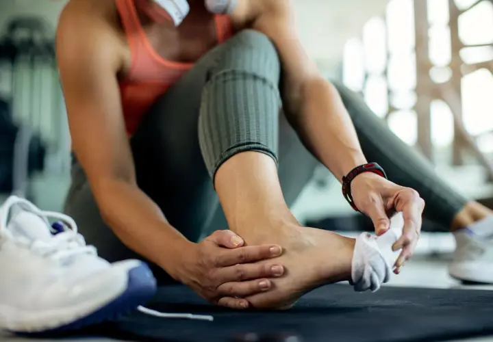 Kobieta w siłowni masuje bolącą kostkę, podejrzewając stan zapalny ścięgna. Obok leżą buty sportowe.
