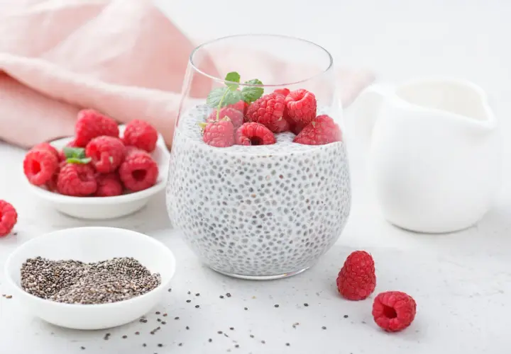 Pyszny deser chia pudding z malinami i miętą, obok miseczka z nasionami chia i świeżymi malinami.