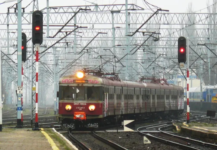 Czerwony pociąg EN57 stoi na torach, sygnalizując czerwonym światłem. Słupy z czerwono-białymi pasami i sygnalizatory kolejowe dźwiękowe wskazują na gotowość do odjazdu.